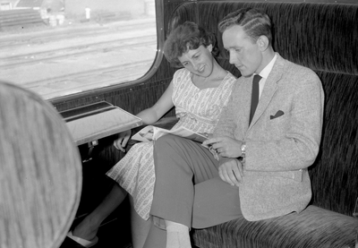 152862 Afbeelding van twee reizigers in een trein, met het tijdschrift Tussen de Rails van de N.S.